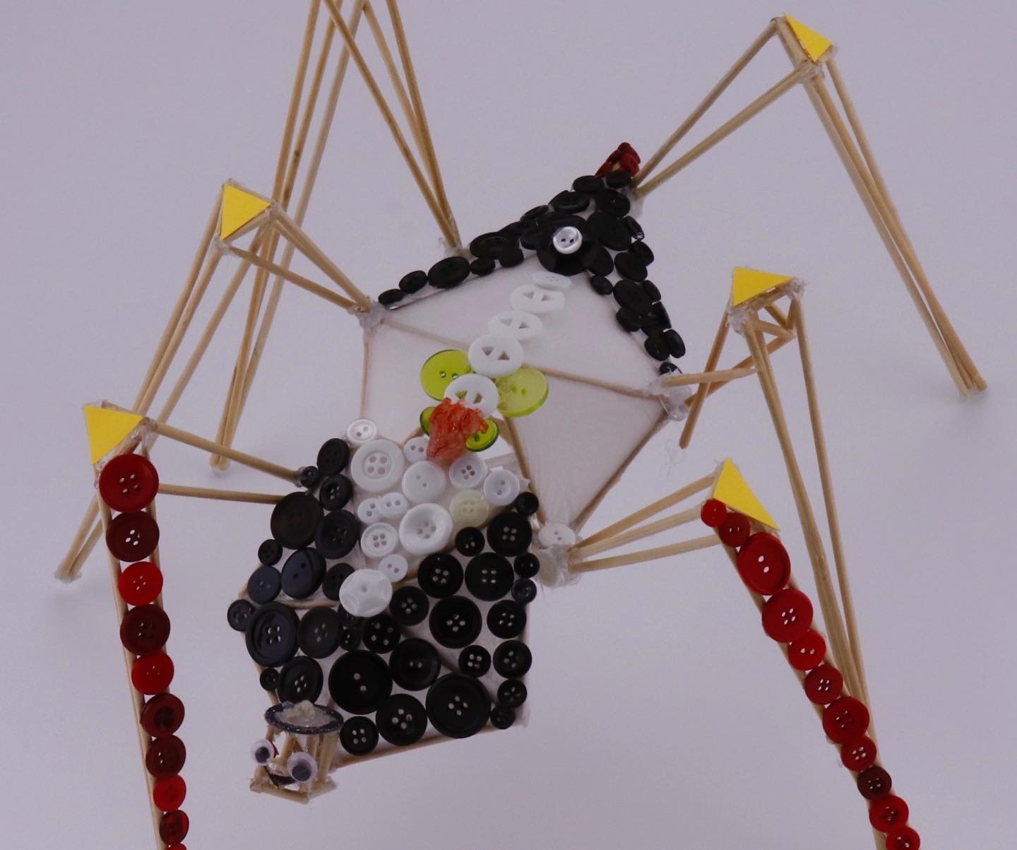 spider scultpure of wood skewers, buttons, two red legs, pattern, googley eyes.