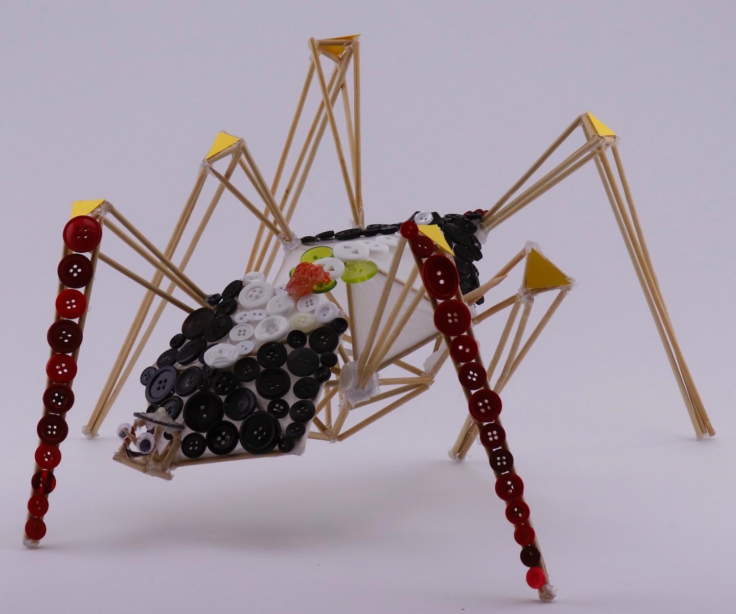 spider scultpure of wood skewers, buttons, two red legs, pattern, googley eyes.